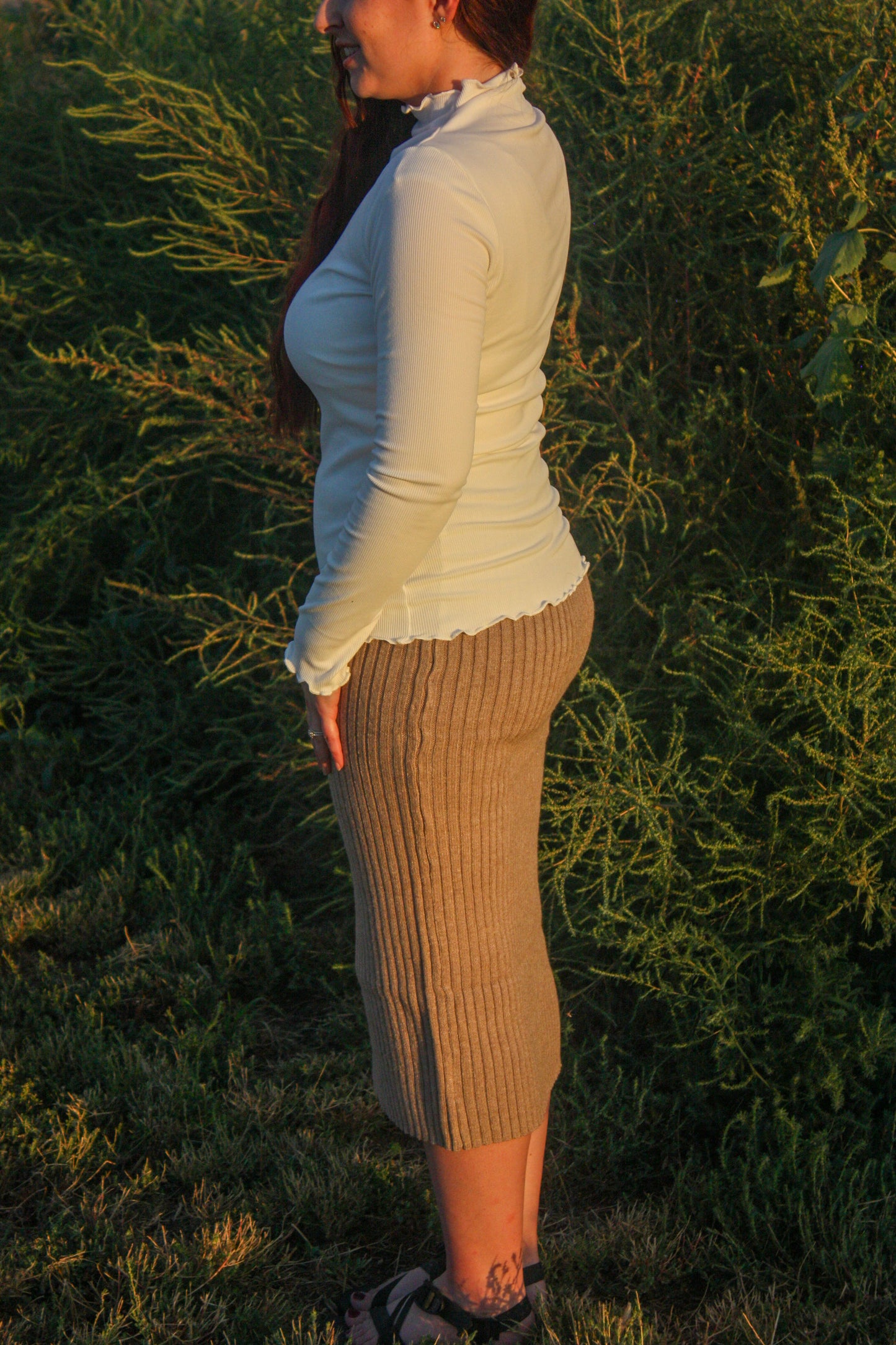 Woman standing in a field with long hair and a white mock neck long sleeve top with merrow edge details and a tan ribbed midi skirt