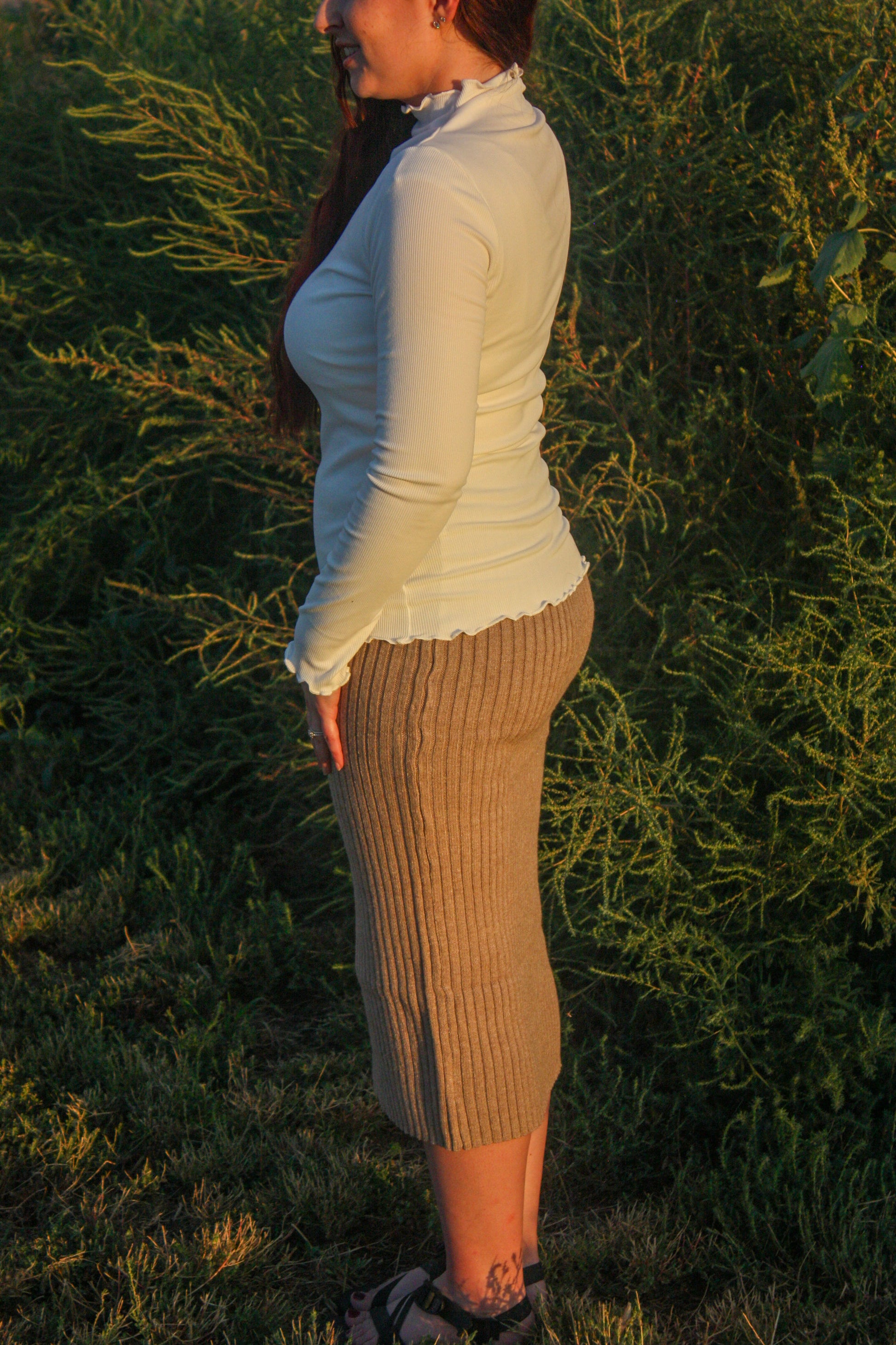 Woman standing in a field with long hair and a white mock neck long sleeve top with merrow edge details and a tan ribbed midi skirt