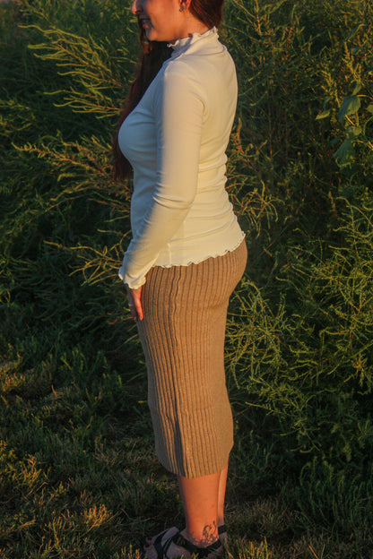 Woman standing in a field with long hair and a white mock neck long sleeve top with merrow edge details and a tan ribbed midi skirt
