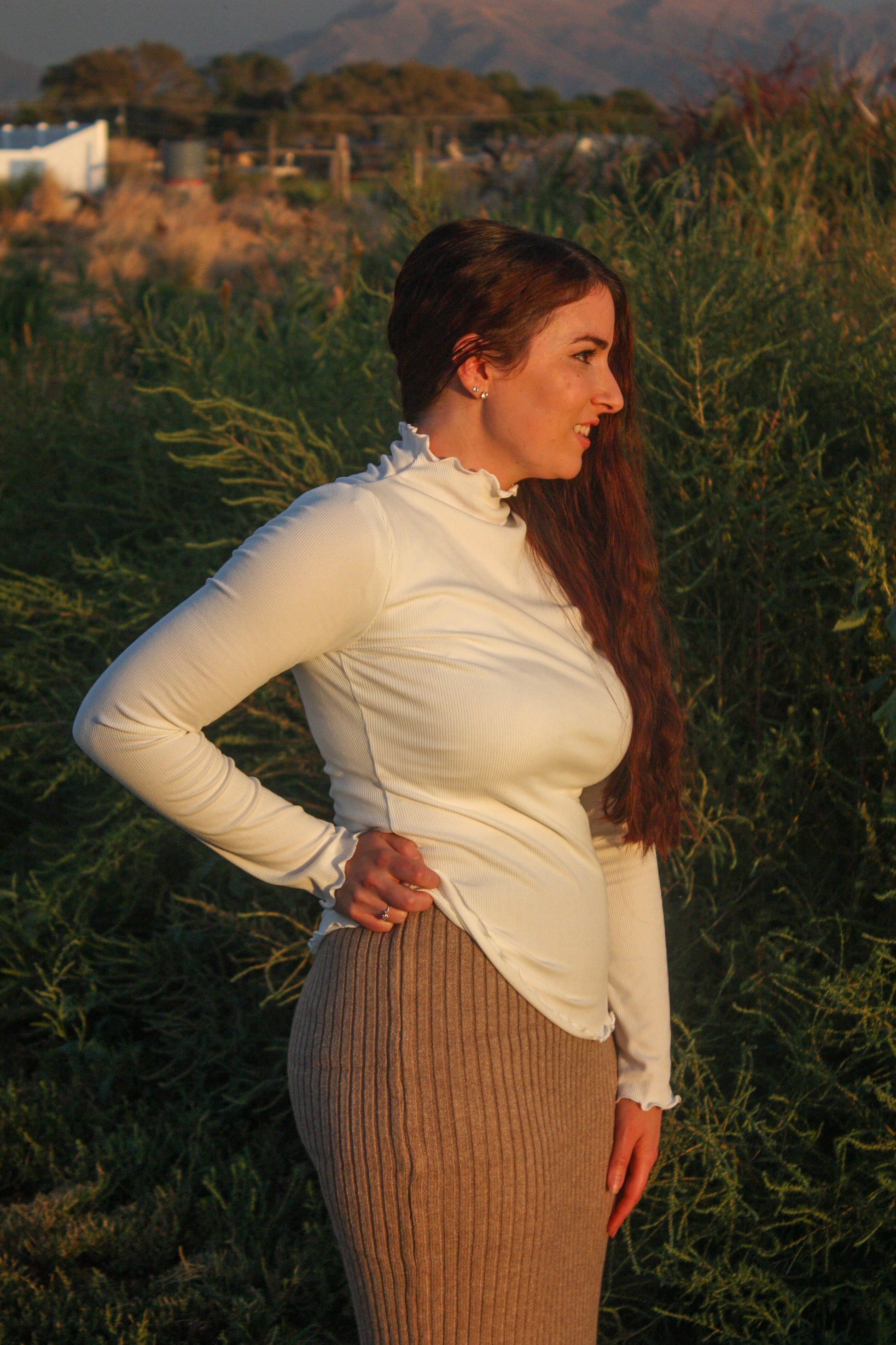 Woman standing in a field with long hair and a white mock neck long sleeve top with merrow edge details and a tan ribbed midi skirt
