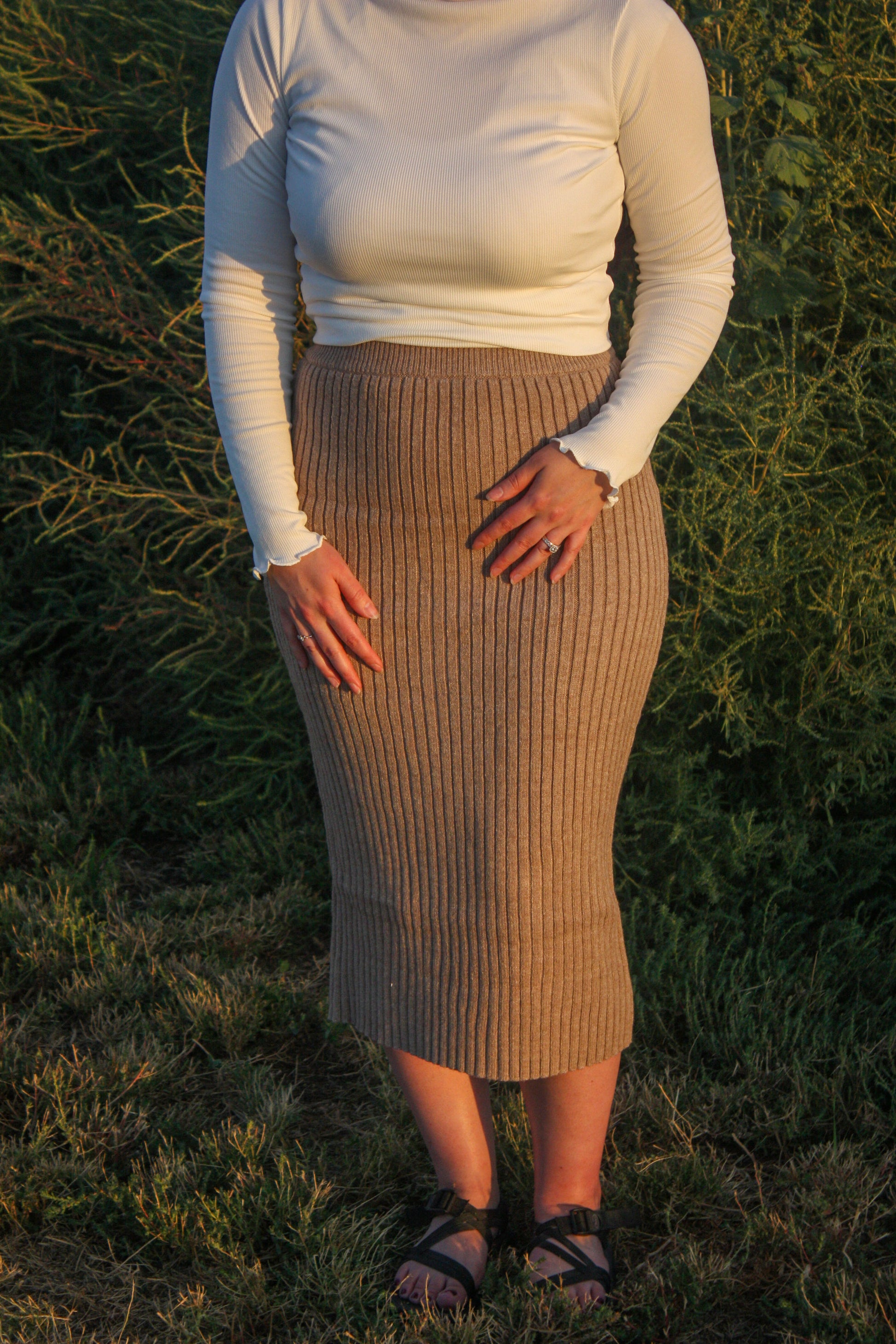 Person wearing a white long-sleeve top and beige ribbed skirt standing in a grassy area.