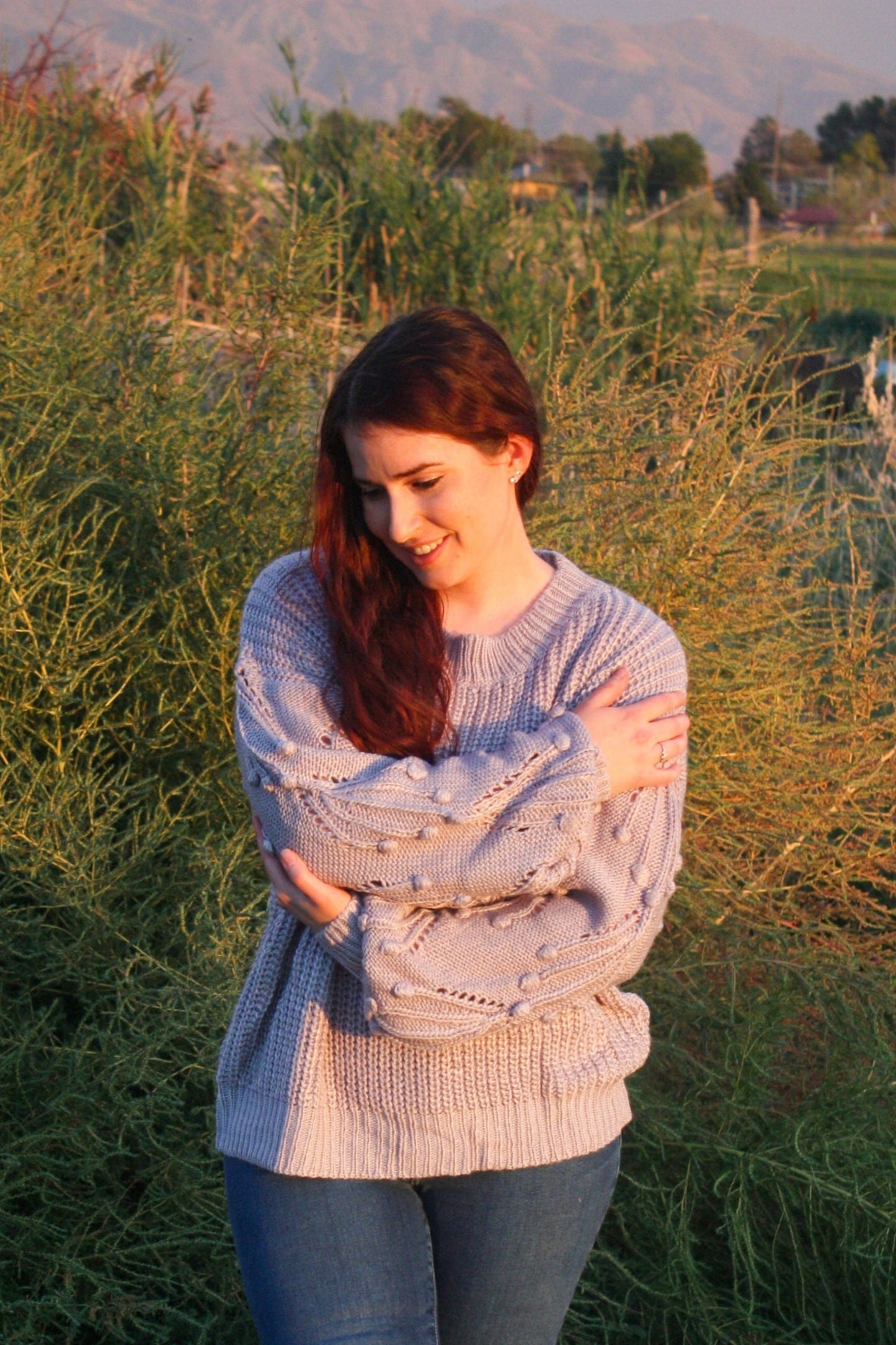 Woman standing in a field with mountains in the background wearing a blue grey sweater with pom details on the balloon sleeves.