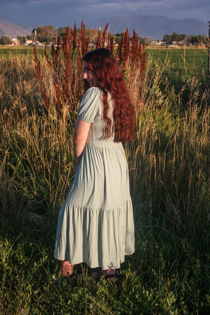 Woman in a light sage green dress standing in a field with mountains in the background showing a side view of the dress. The dress has puff sleeves, short in length. The dress is a midi dress that has tiers in the skirt.