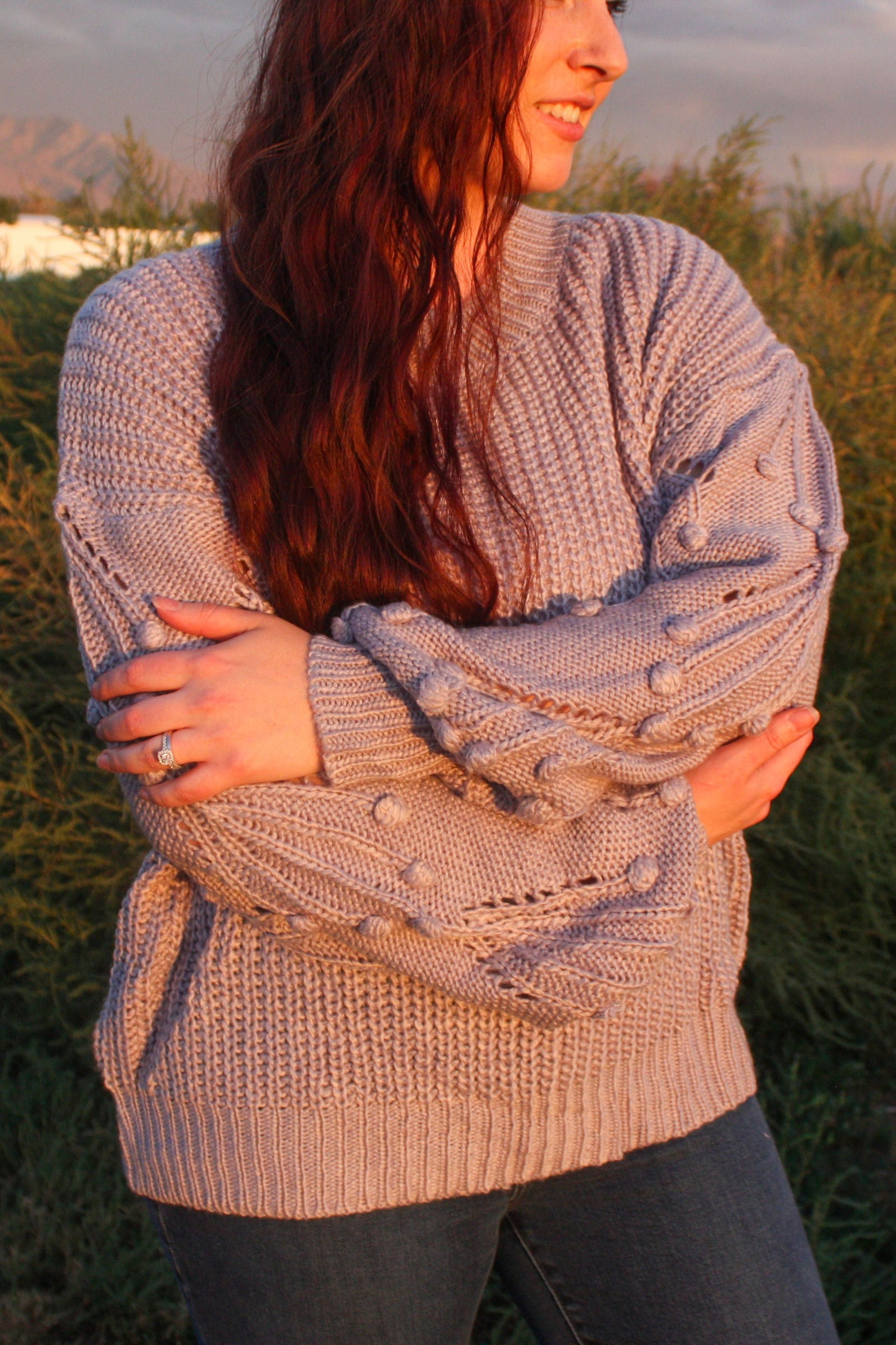 Woman standing in a field with mountains in the background wearing a blue grey sweater with pom details on the balloon sleeves.