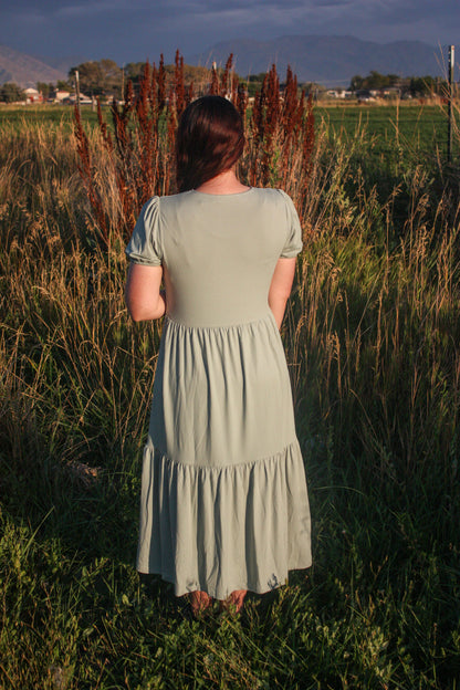 Woman in a light sage green dress standing in a field with mountains in the background showing a back view of the dress. The dress has puff sleeves, short in length. The dress is a midi dress that has tiers in the skirt.