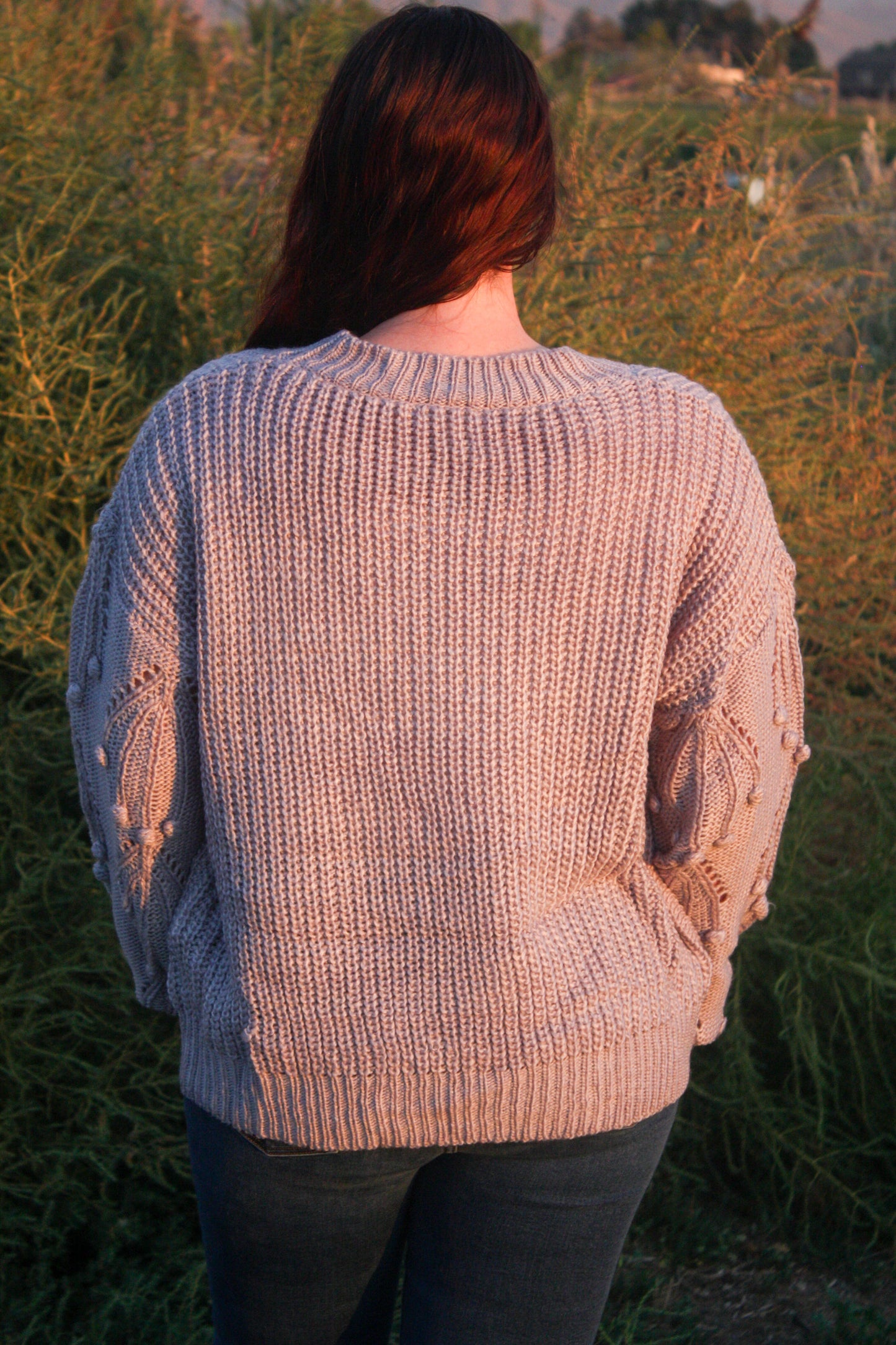 Woman standing in a field with mountains in the background wearing a blue grey sweater with pom details on the balloon sleeves.