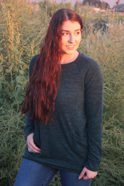 Woman standing in a field with long hair and a hunter green knit long sleeve top with front pockets.