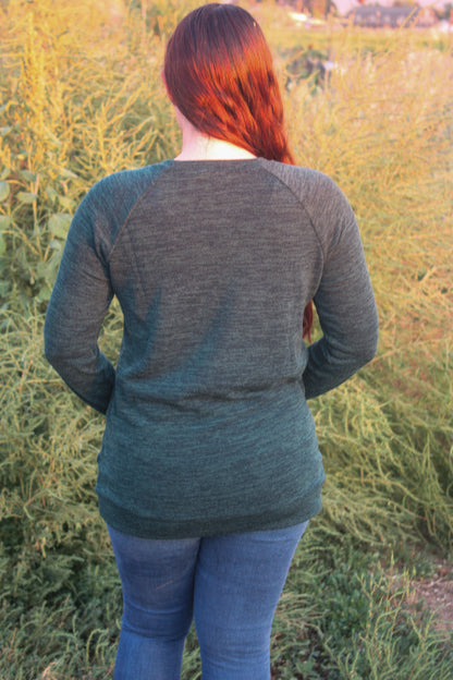 Woman standing in a field with long hair and a hunter green knit long sleeve top with front pockets.