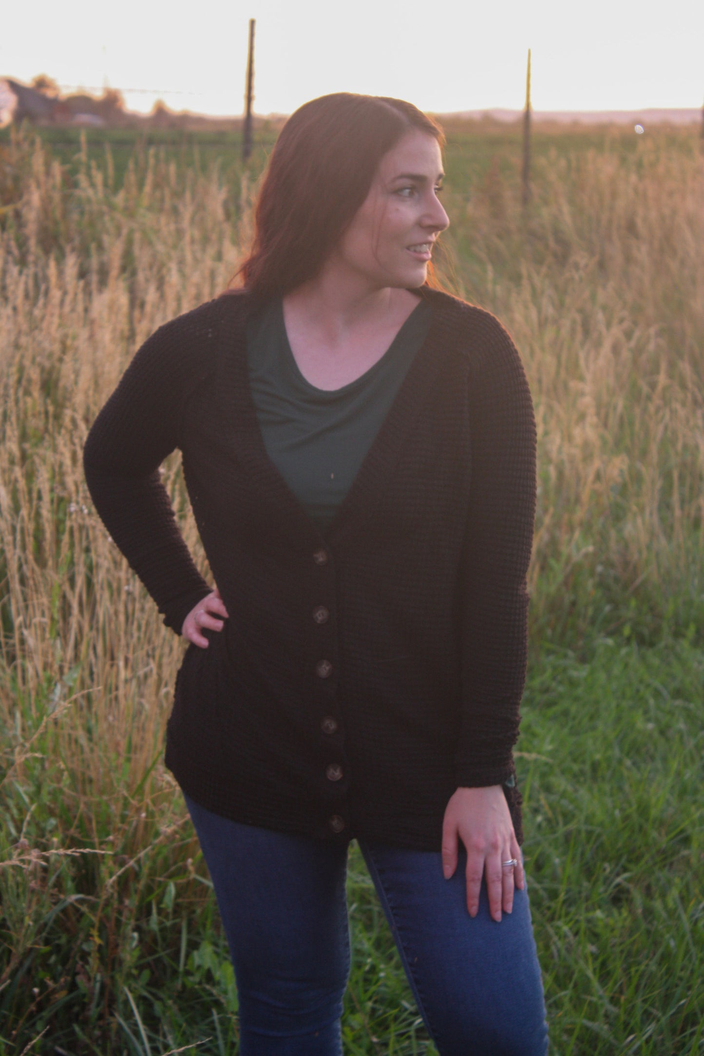 Woman standing in a field wearing a black cardigan and blue jeans.