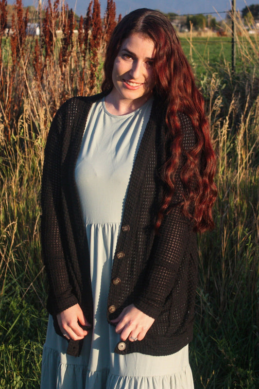 Woman with long red hair standing in a field of tall grass and plants wearing a light green dress with tiers and a black cardigan with brown buttons.