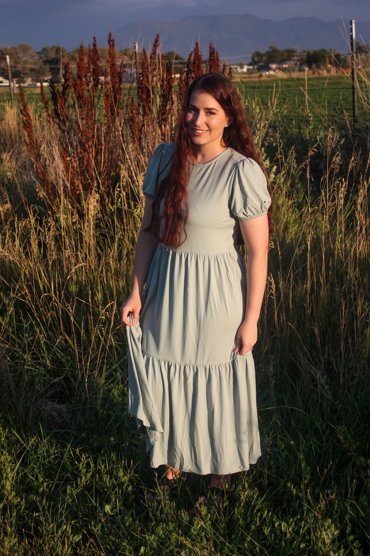 Woman in a light sage green dress standing in a field with mountains in the background. The dress has puff sleeves, short in length. The dress is a midi dress that has tiers in the skirt.