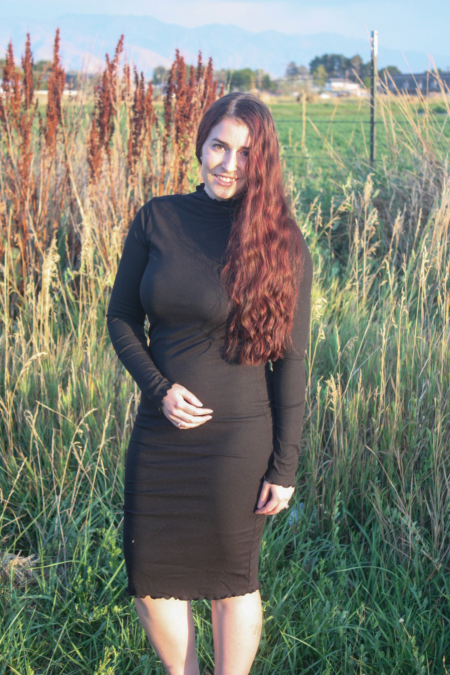 Woman in a midi length form fitting long sleeve black dress with a mock neck and merrow edge details standing in a field with tall grass and plants.