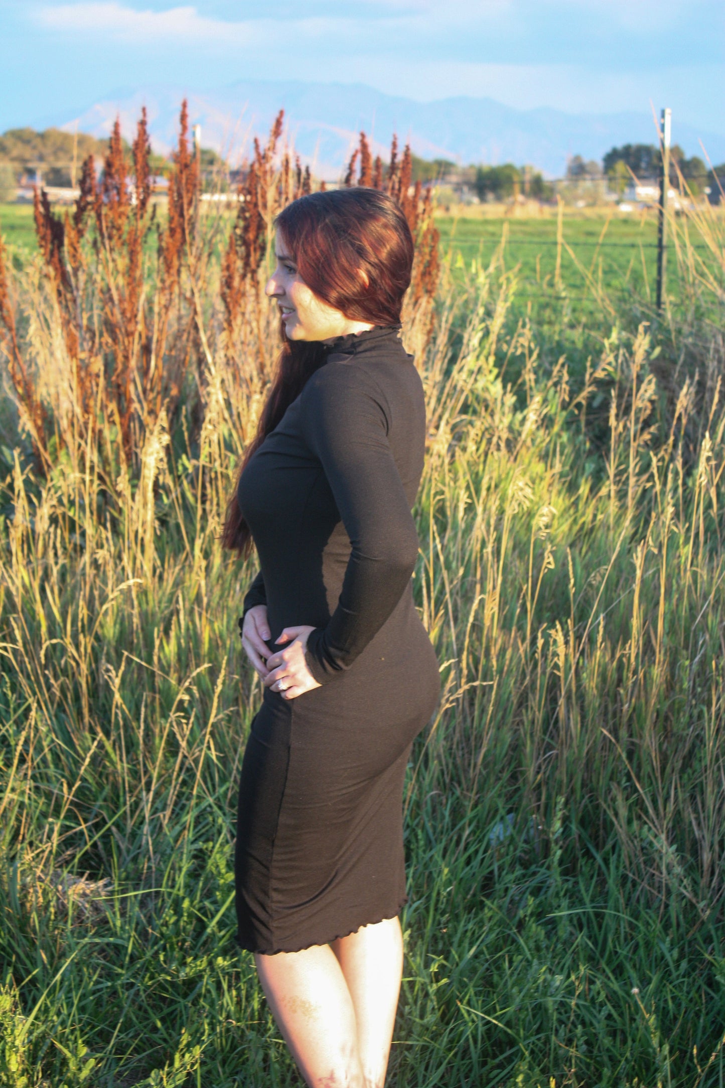 Woman in a midi length form fitting long sleeve black dress with a mock neck and merrow edge details standing in a field with tall grass and plants.