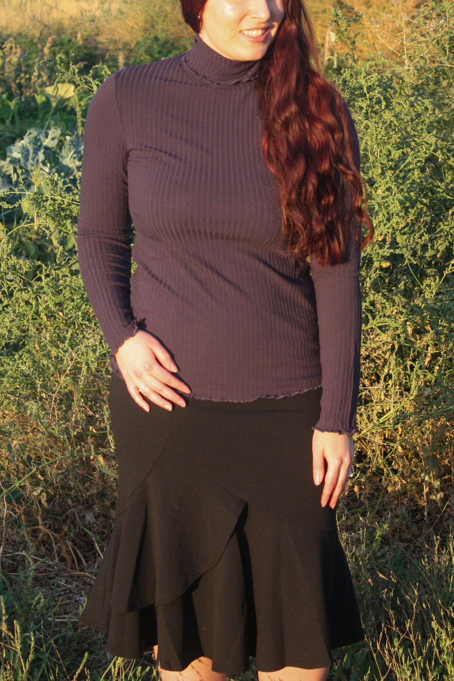 Woman wearing a navy ribbed mock neck, long-sleeve top with lettuce hem detailing and black skirt standing in a natural setting.