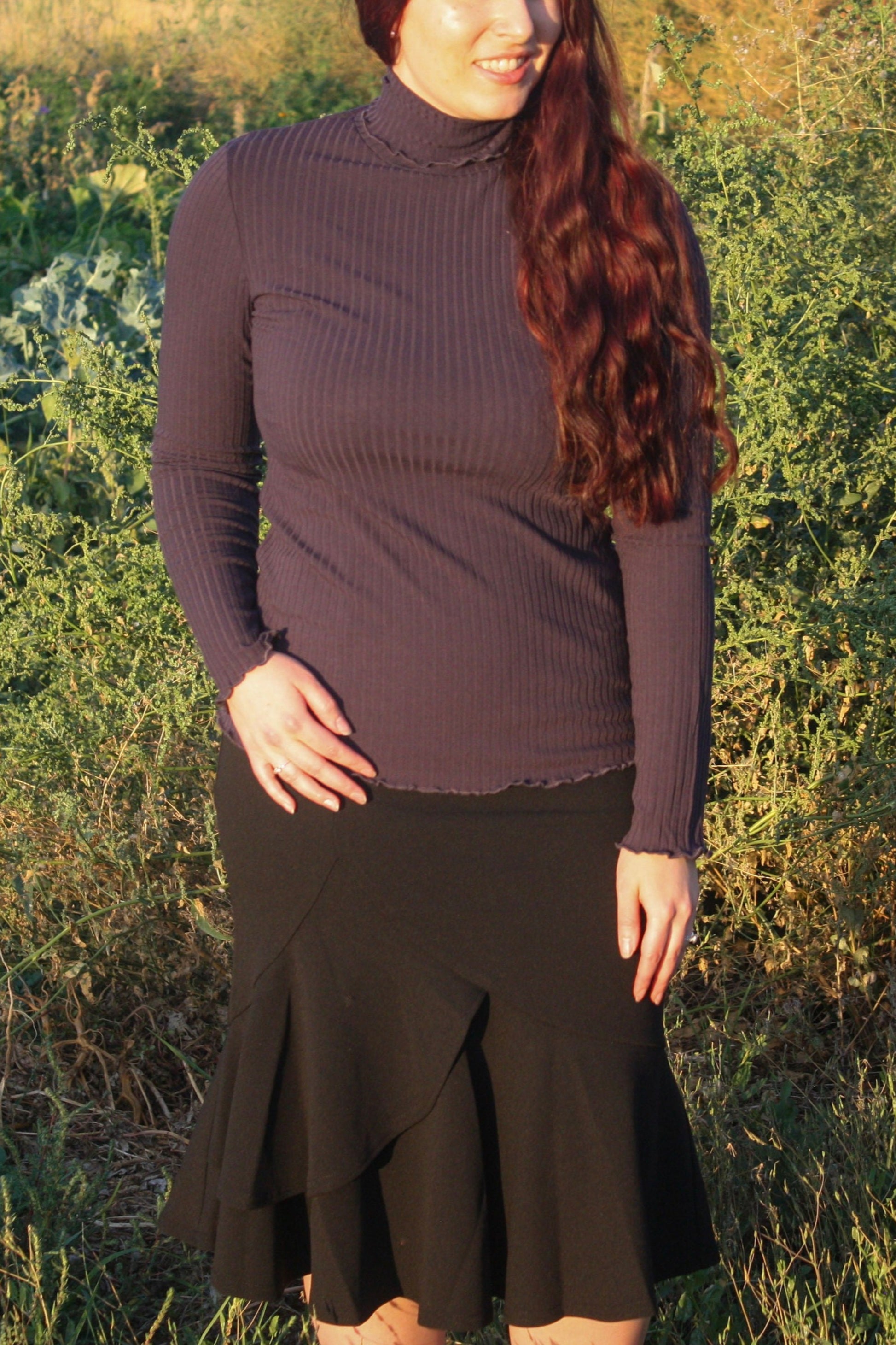Woman wearing a navy ribbed mock neck, long-sleeve top with lettuce hem detailing and black skirt standing in a natural setting.