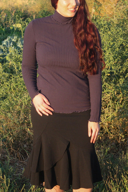 Woman wearing a navy ribbed mock neck, long-sleeve top with lettuce hem detailing and black skirt standing in a natural setting.