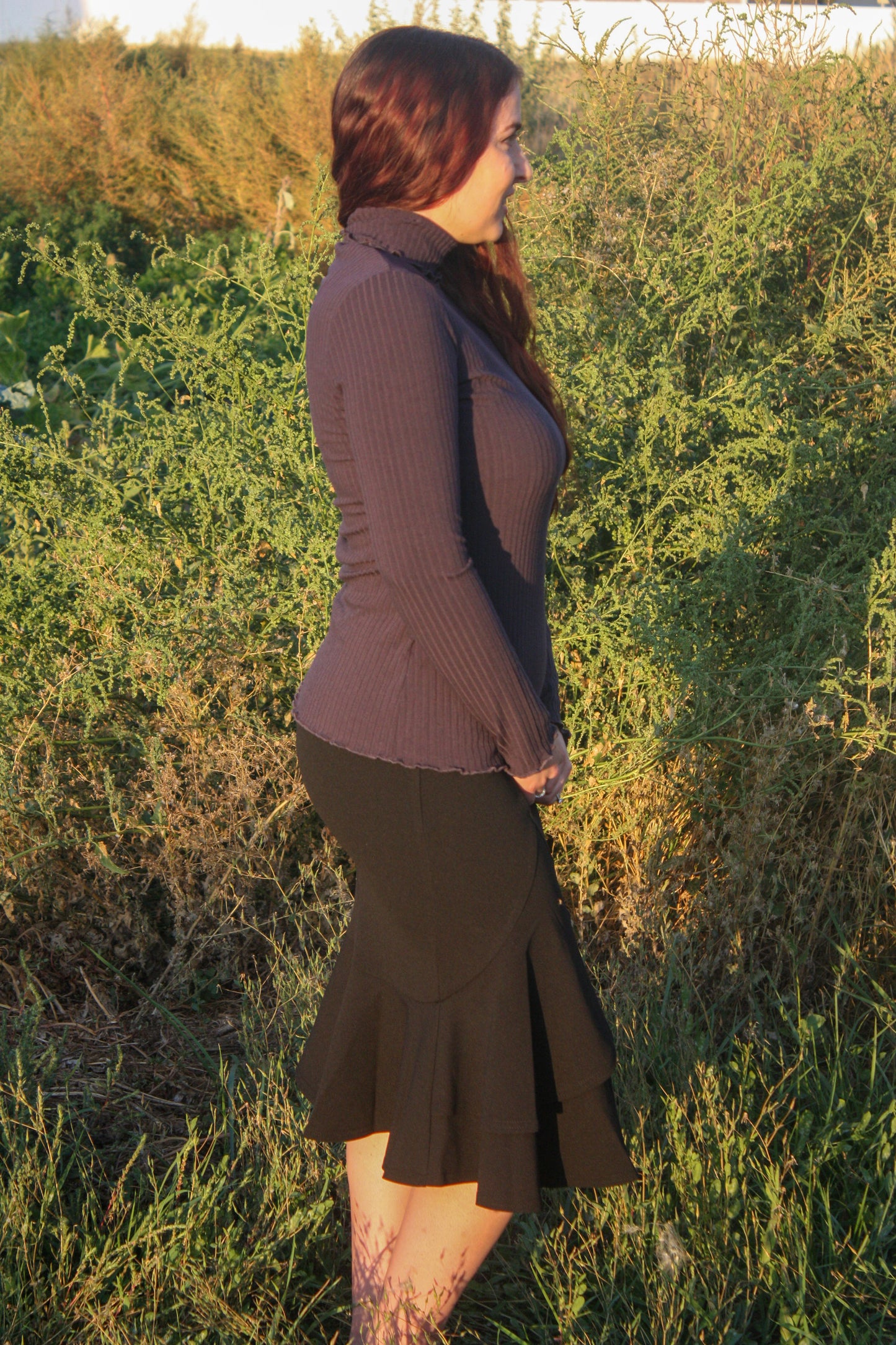 Woman wearing a navy ribbed mock neck, long-sleeve top with lettuce hem detailing and black skirt standing in a natural setting.