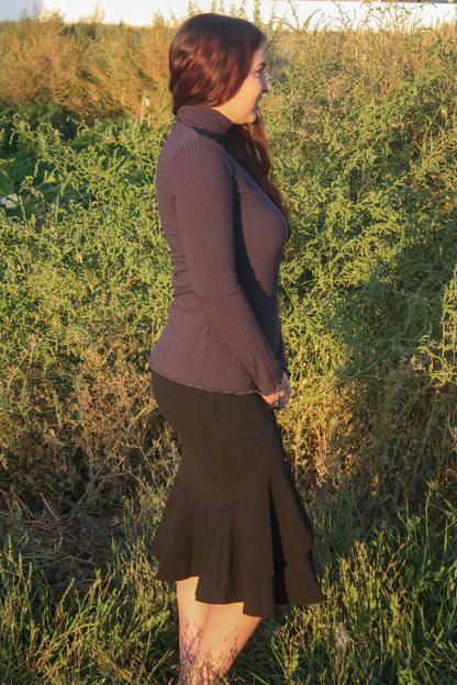 Woman wearing a navy ribbed mock neck, long-sleeve top with lettuce hem detailing and black skirt standing in a natural setting.