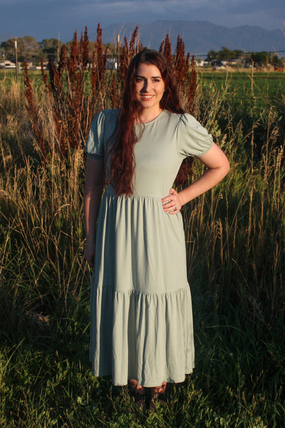 Woman in a light sage green dress standing in a field with mountains in the background. The dress has puff sleeves, short in length. The dress is a midi dress that has tiers in the skirt.