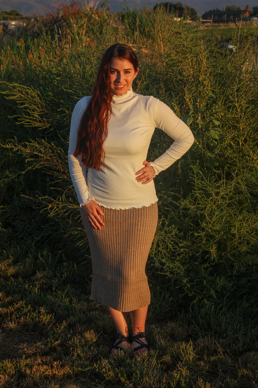 Woman standing in a field with long hair and a white mock neck long sleeve top with merrow edge details and a tan ribbed midi skirt.